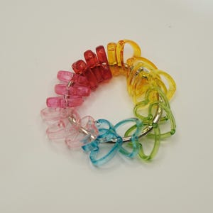 May include: A colorful hair tie featuring a rainbow of hues, including pink, red, yellow, green, and blue. The hair tie is made of clear plastic and is arranged in a circular shape, with each color segment connected by a silver metal ring.