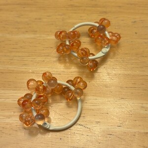 May include: Two circular white plastic rings adorned with clusters of small, translucent orange spherical beads. Each ring features a hinged mechanism for opening and closing, with the beads attached around the outer circumference of the white frame.
