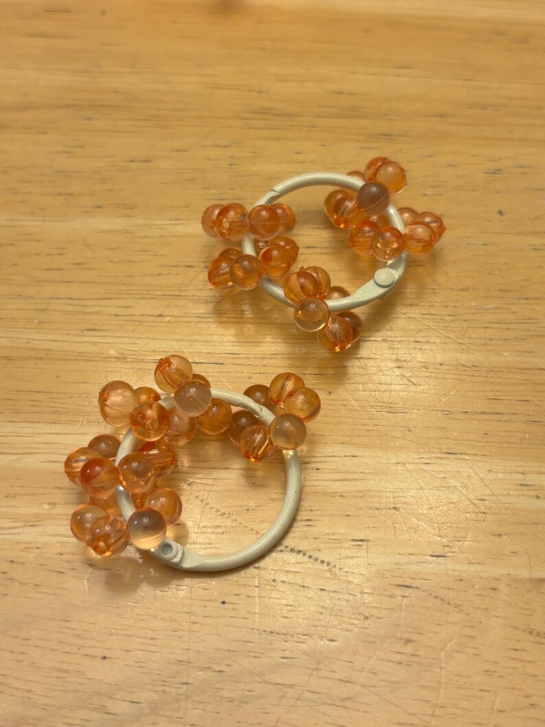 May include: Two circular white plastic rings adorned with clusters of small, translucent orange spherical beads. Each ring features a hinged mechanism for opening and closing, with the beads attached around the outer circumference of the white frame.