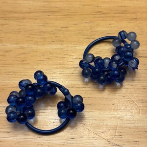 May include: A pair of circular hair ties featuring clusters of translucent dark blue spherical beads attached to a solid dark blue elastic band. The beads vary slightly in shade and transparency, creating a textured, rounded appearance on each hair accessory.