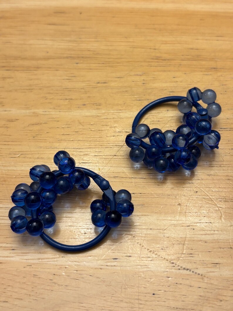 May include: A pair of circular hair ties featuring clusters of translucent dark blue spherical beads attached to a solid dark blue elastic band. The beads vary slightly in shade and transparency, creating a textured, rounded appearance on each hair accessory.