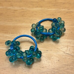 May include: A pair of hoop earrings featuring a bright blue circular frame adorned with numerous translucent teal beads. The beads are clustered around the circumference of each hoop, creating a textured, spherical appearance on the blue metal base.