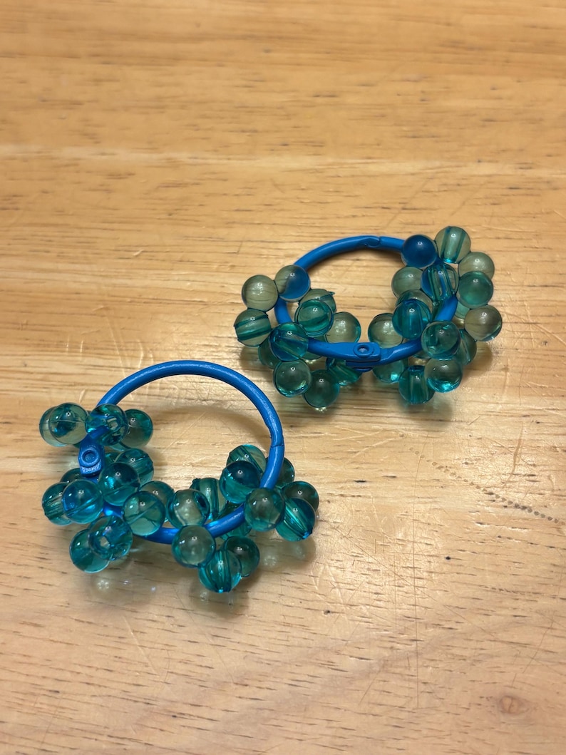 May include: A pair of hoop earrings featuring a bright blue circular frame adorned with numerous translucent teal beads. The beads are clustered around the circumference of each hoop, creating a textured, spherical appearance on the blue metal base.