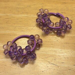 May include: A pair of hoop earrings featuring a purple circular frame adorned with clusters of translucent, light purple spherical beads. The beads are arranged in small groups around the circumference of each hoop, creating a textured, multi-dimensional appearance with a glossy finish.