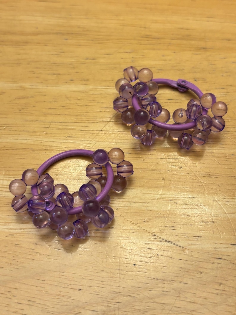 May include: A pair of hoop earrings featuring a purple circular frame adorned with clusters of translucent, light purple spherical beads. The beads are arranged in small groups around the circumference of each hoop, creating a textured, multi-dimensional appearance with a glossy finish.