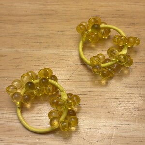 May include: A pair of circular yellow hair ties, each featuring clusters of translucent yellow spherical beads attached to the band. The beads are arranged in small groups around the circumference of the yellow elastic hair accessory.