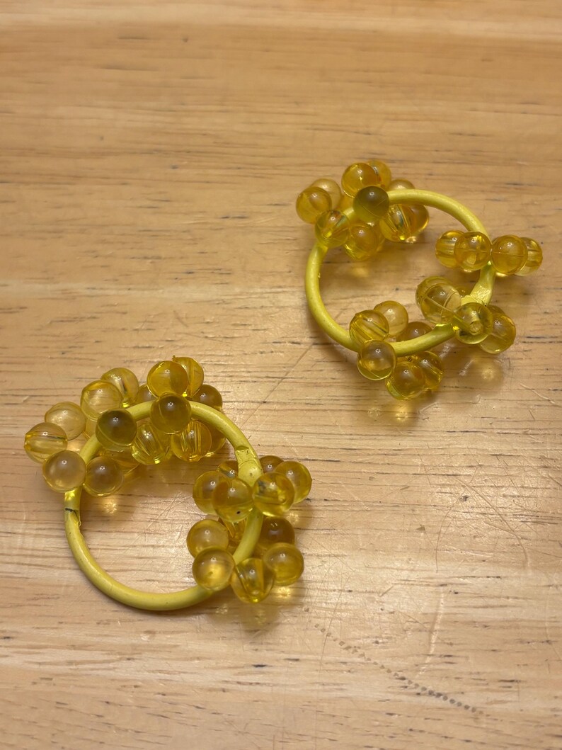May include: A pair of circular yellow hair ties, each featuring clusters of translucent yellow spherical beads attached to the band. The beads are arranged in small groups around the circumference of the yellow elastic hair accessory.