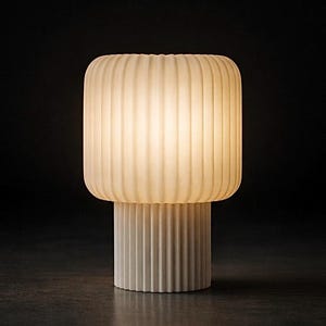 Könnte beinhalten: Moderne Tischlampe in Off-White mit geripptem, rechteckigem Schirm auf zylindrischem Sockel. Die Lampe strahlt ein weiches, warmes Licht vor dunklem Hintergrund aus. Minimalistisches und elegantes Design für verschiedene Einrichtungsstile.