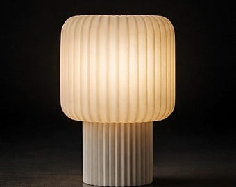 Puede incluir: Lámpara de mesa moderna en blanco roto, con pantalla rectangular y acanalada sobre una base cilíndrica. La lámpara emite una luz suave y cálida sobre un fondo oscuro. Diseño minimalista y elegante, adecuado para varios interiores.