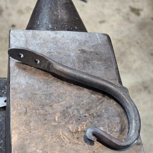 May include: A hand-forged metal hook with a curved design, featuring two holes on the flat end for mounting. The hook is a dark gray color and rests on a metal anvil, showcasing the craftsmanship of the blacksmith.