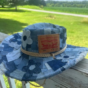May include: A patchwork denim bucket hat featuring various shades of blue fabric squares and stitched flower appliques. A tan leather Levi Strauss & Co. patch is centered on the front, and a braided tan rope cord encircles the crown base.
