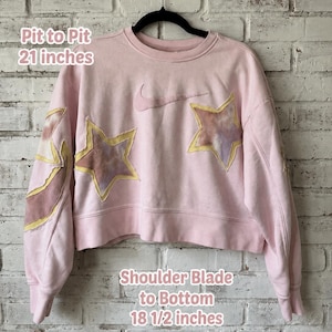 May include: A light pink cropped sweatshirt featuring a Nike swoosh logo and star-shaped patches with a tie-dye pattern. Text on the image indicates a pit to pit measurement of 21 inches and a shoulder blade to bottom length of 18 1/2 inches.