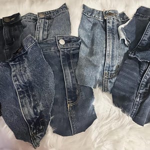 May include: A collection of six denim jean scraps in various shades of blue, including black, light blue, and dark blue. The scraps are arranged on a white fluffy surface.