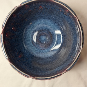 May include: A handmade ceramic bowl with a deep blue glaze and a scalloped rim. The bowl's interior has a gradient of blues, with hints of purple and red accents. The rim is edged with a lighter color, creating a contrast.