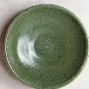 May include: A handmade, round, green ceramic bowl with a textured surface. The bowl has a slightly raised rim and a smooth, glossy interior. The color is a muted, earthy green, perfect for serving salads or side dishes.
