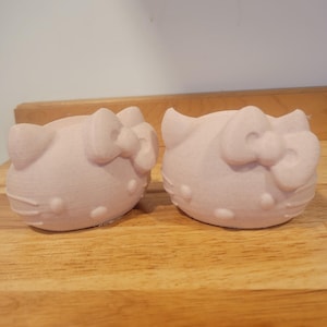 May include: Two light pink, 3D-printed Hello Kitty head-shaped objects. Each has a bow and facial features. They are sitting on a wooden surface. The objects are likely decorative or functional items.
