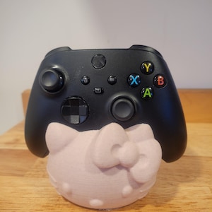 Può includere: Controller Xbox nero appoggiato su un supporto rosa chiaro a forma di Hello Kitty. Il controller ha pulsanti colorati e controlli direzionali. Il supporto è stampato in 3D, aggiungendo un tocco giocoso alla configurazione di gioco. Circa 10 cm di altezza.