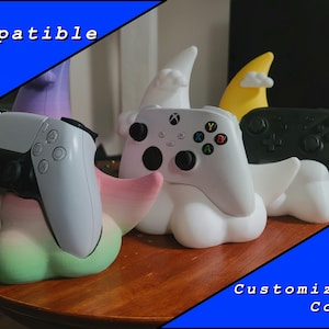 Puede incluir: Soportes para mandos de videojuegos con forma de nube, cada uno sosteniendo un mando diferente. Un soporte sostiene un mando PS5 blanco, otro un mando Xbox y un tercero un mando negro. Los soportes son blancos con acentos coloridos y el texto "PS5 Compatible" y "Customizable Colors".