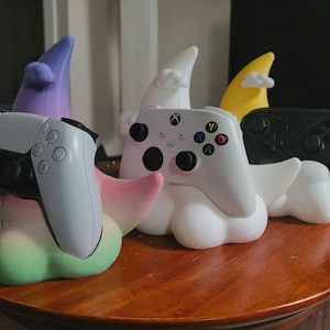 以下が含まれることがあります： 雲と月の形をした、カラフルなコントローラー用スタンドのコレクション。各スタンドには、白いPlayStation 5コントローラー、白いXboxコントローラー、黒いコントローラーなど、さまざまなゲームコントローラーが置かれています。