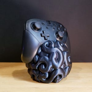 May include: A black Nintendo Switch Pro Controller rests on a 3D-printed brain-shaped stand. The controller features buttons, joysticks, and a directional pad. The stand is a dark, textured sculpture, providing a unique aesthetic for gamers.