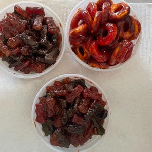 May include: Three plastic containers filled with assorted candies. One container holds dark red and brown rectangular candies. Another contains red, ring-shaped candies. The third container has a mix of red and brown candies. All are on a white surface.