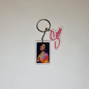 May include: A keychain featuring a rectangular pendant with a portrait of a woman wearing a colorful top. The keychain also includes a pink, cursive "Selena" charm and a silver key ring.
