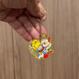 May include: A glittery acrylic keychain featuring a cartoon illustration of a character with yellow hair and a rainbow-colored outfit alongside a small white creature with rainbow-striped legs, attached to a silver-toned metal split ring and chain.