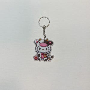 May include: A keychain featuring a cartoon cow character with a white and black cow pattern, wearing a pink bow and holding a coffee cup. The keychain has a silver-colored ring and chain, and is decorated with rhinestones.