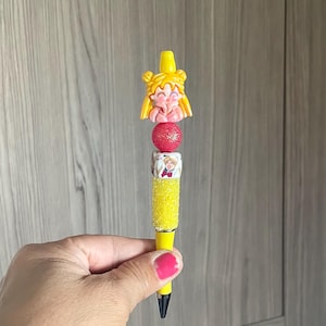 May include: A yellow pen with a decorative topper featuring a cartoon character with blonde hair. The pen has a pink glitter bead and a section with a cartoon image. The pen is held in a hand with pink nail polish.