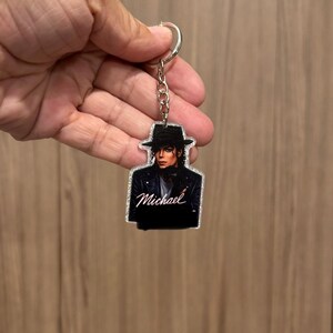 May include: A glittery acrylic keychain featuring a printed image of a person wearing a black fedora and a black leather jacket. The name Michael is printed in white cursive script across the chest area of the figure on the keychain.