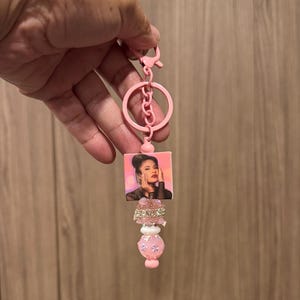May include: Pink keychain featuring a square image of a woman with red lipstick. The keychain has a pink ring, a chain, and decorative pink and white beads. A stylish accessory.