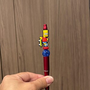 May include: A red pen with a Bart Simpson figure on top. The pen has a blue and red bead design. The Bart Simpson figure is holding a skateboard. The pen is held in a hand.
