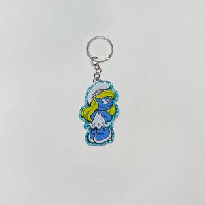 May include: A Smurfette keychain featuring a blue Smurfette character with yellow hair and a white dress. The keychain has a silver ring and chain. The Smurfette design is outlined with blue glitter.