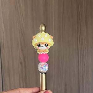May include: A gold stylus pen with a mushroom-shaped topper. The topper features a yellow mushroom cap with white polka dots, a cute cartoon face, and a pink ball. Below is a clear ball with colorful glitter.