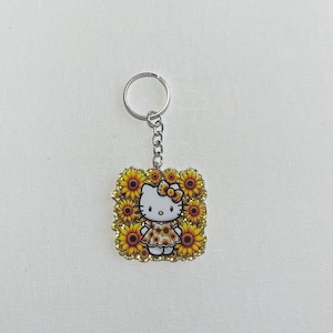 May include: A keychain featuring a cartoon cat character surrounded by sunflowers. The cat wears a sunflower-patterned dress and bow. The keychain has a silver-colored chain and ring.