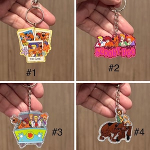 May include: Four acrylic keychains featuring Scooby-Doo characters. Keychain 1 shows three polaroid photos labeled THE GANG. Keychain 2 has pink glitter and the text SCOOBY-DOO. Keychain 3 depicts the Mystery Machine van. Keychain 4 shows Scooby and Shaggy.