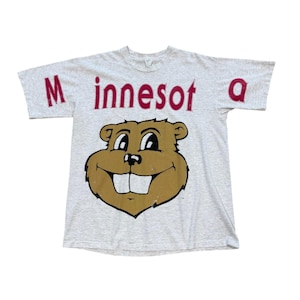Könnte beinhalten: Ein hellgrau meliertes Kurzarm-T-Shirt mit dem roten Schriftzug "Minnesot a" auf der Brust. Unter dem Text befindet sich eine große, zentrierte Grafik eines braunen Comic-Gopher-Kopfes mit schwarzen Umrissen, weißen Zähnen und einem Lächeln.