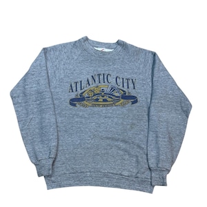 May include: A heather gray long-sleeve crewneck sweatshirt featuring a graphic print on the chest. The design includes the text "ATLANTIC CITY" above an illustration of dice, a roulette wheel, and chips, with "NEW JERSEY" below.