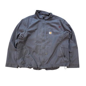 May include: A grey Carhartt zip-up jacket featuring a stand-up collar, a vertical zipped chest pocket, and two lower front pockets. The jacket displays a small rectangular Carhartt brand logo patch on the chest and a small red kangaroo embroidery.