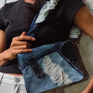 以下が含まれることがあります： 再利用された青いデニム生地で作られたショルダーバッグ。パッチワークデザイン、ほつれたエッジ、使い古された質感、幅広のギャザー入りデニムストラップが特徴です。バッグにはダークブルーのデニムパネルと、白い糸が見える中央部分が含まれています。