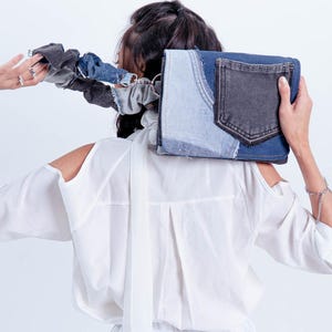 以下が含まれることがあります： 濃いグレーのポケットと薄いブルーのデニムアクセントが付いたデニムクラッチ。クラッチはリサイクルデニムで作られており、お揃いのデニムシュシュが付いています。白いシャツを着た人が持っています。