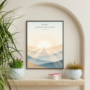 May include: Framed art print featuring a watercolor landscape with layered blue and tan hills, and a bright sunburst. The text "Be Still, and know that I am God" is at the top. The print is displayed on a wooden shelf with plants.