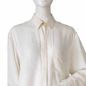 Puede incluir: Camisa crema de manga larga con cuello clásico y un bolsillo en el pecho. La camisa tiene un botón dorado en el cuello y está hecha de una tela ligera y fluida.