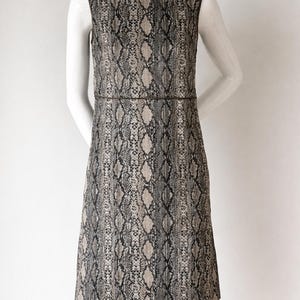 May include: Sleeveless sheath dress with a snakeskin pattern in shades of gray, black, and beige. The dress has a round neckline and a fitted waist. The dress is displayed on a mannequin.
