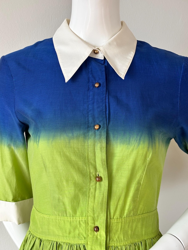 Puede incluir: Vestido degradado azul y verde con cuello blanco y botones. El vestido presenta mangas cortas con pu&ntilde;os blancos y una falda plisada. El degradado de color pasa del azul al verde, creando un dise&ntilde;o vibrante y llamativo. El vestido mide unos 100 cm de largo.