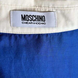 Puede incluir: Primer plano de una prenda azul con cuello blanco. Una etiqueta rectangular con el texto "MOSCHINO CHEAPANDCHIC" est&aacute; cosida a la prenda. El fondo es de color verde brillante.