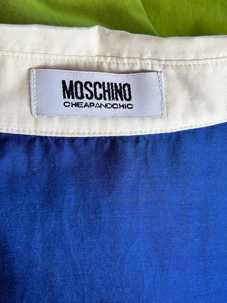 Puede incluir: Primer plano de una prenda azul con cuello blanco. Una etiqueta rectangular con el texto "MOSCHINO CHEAPANDCHIC" est&aacute; cosida a la prenda. El fondo es de color verde brillante.
