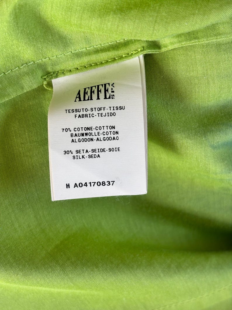Puede incluir: Primer plano de una etiqueta blanca con el nombre de la marca "AEFFE" impreso, adherida a una tela verde brillante. La etiqueta proporciona detalles de la composici&oacute;n de la tela en varios idiomas.