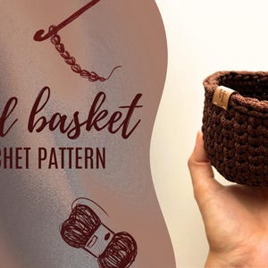 Puede incluir: Una cesta redonda de ganchillo marrón oscuro, sostenida en una mano. La cesta tiene forma cilíndrica y una superficie texturizada. El texto "Round basket CROCHET PATTERN" se muestra en el lado izquierdo de la imagen.