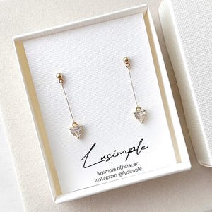 May include: A pair of gold-tone drop earrings featuring a small stud top and a dangling heart-shaped clear stone at the end of a thin chain. The earrings are displayed on a white card printed with the text Lusimple, lusimple.official.ec, and Instagram @lusimple_.